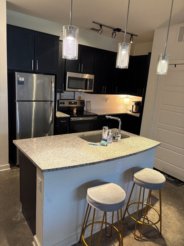 A modern kitchen with a stainless steel refrigerator, a microwave, and a double oven. There are two bar stools with cushioned seats and gold legs.