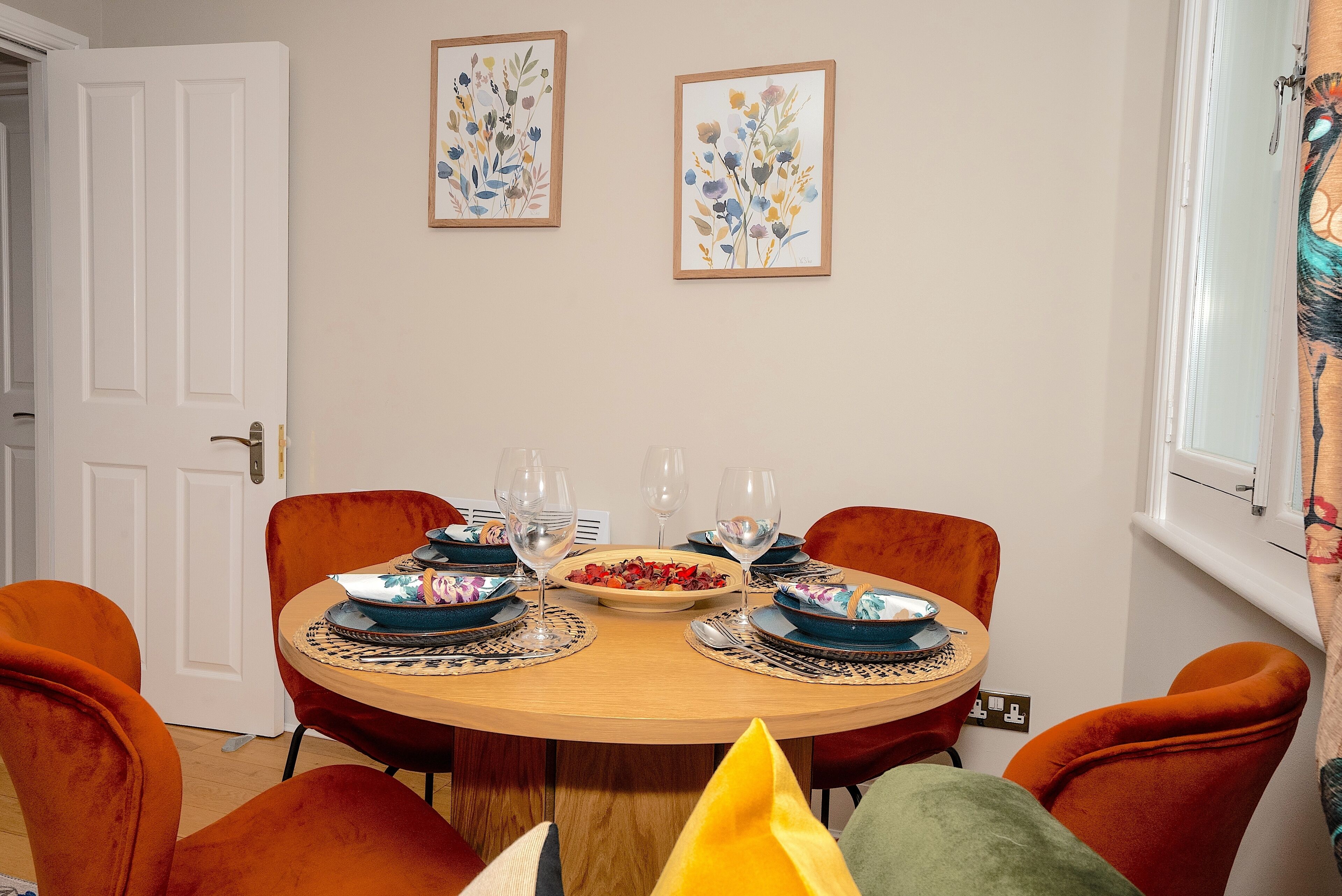 Une salle à manger avec une table ronde dressée pour quatre personnes, agrémentée de chaises orange, de vaisselle bleue et blanche à motifs, et d’un saladier rempli de fruits.