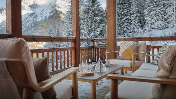 Eine Holzterrasse mit Blick auf schneebedeckte Berge und Bäume. Zwei Holzstühle und ein Tisch mit Gläsern und einem Teekessel darauf.