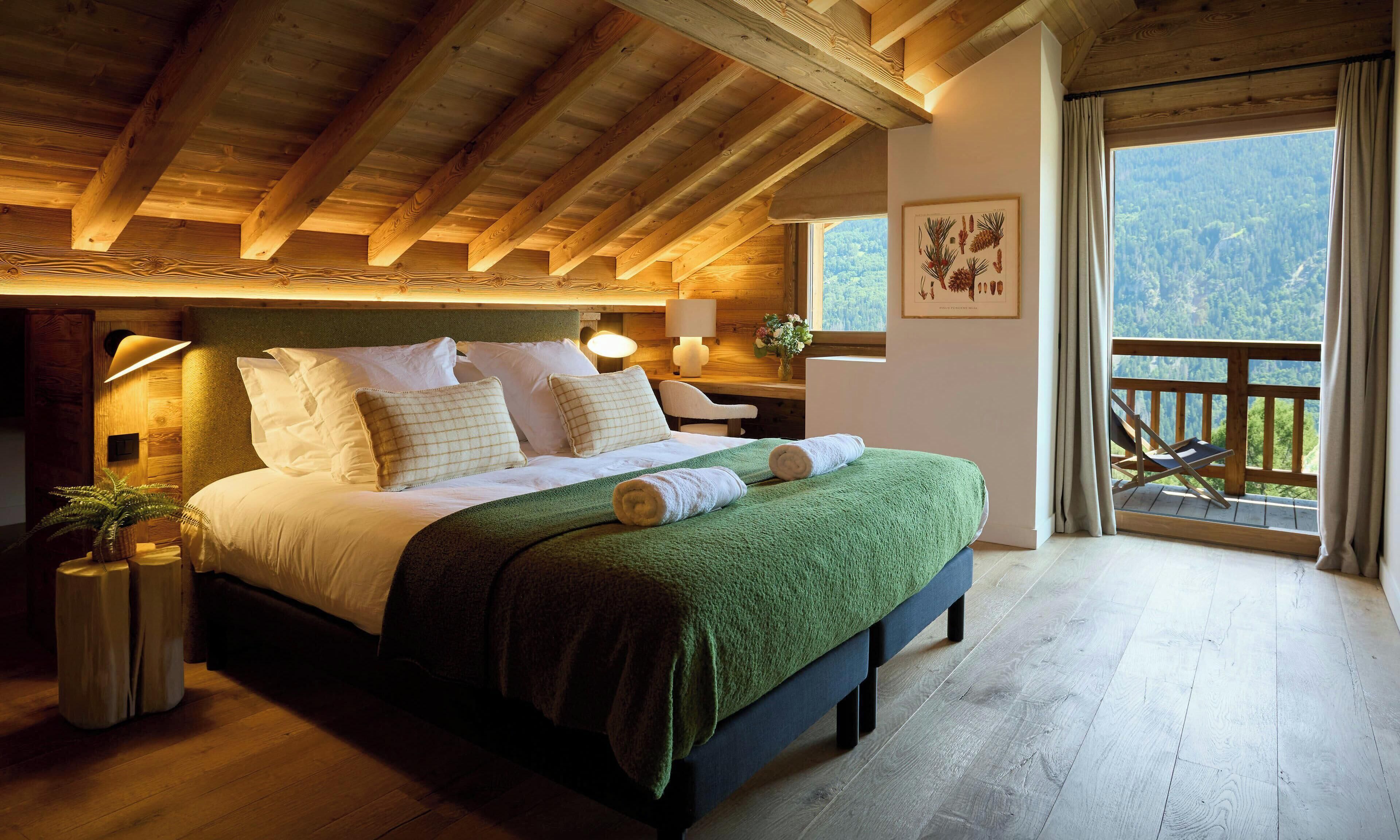 Un dormitorio con techo de madera, una cama con ropa blanca, una manta verde y vistas a un bosque a través de una ventana.