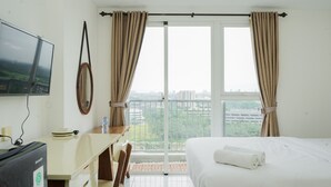 A hotel room with a bed, a desk, a chair, a TV, and a view of the city through large windows. - Simple And Warm Studio At Casa De Parco Apartment (Pagedangan)