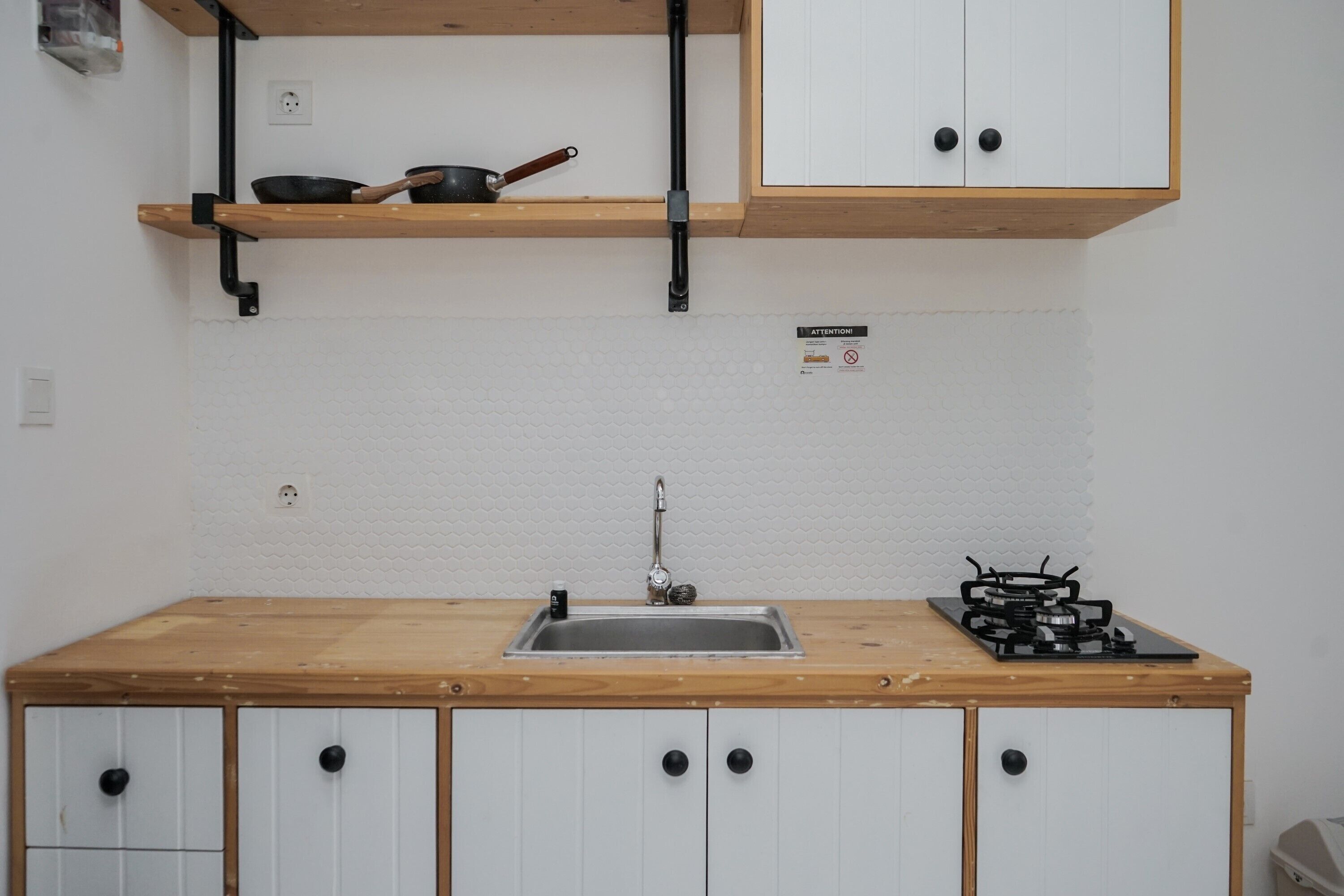 A kitchen with a wooden countertop, a sink, cabinets, a stove, and a shelf with a frying pan.