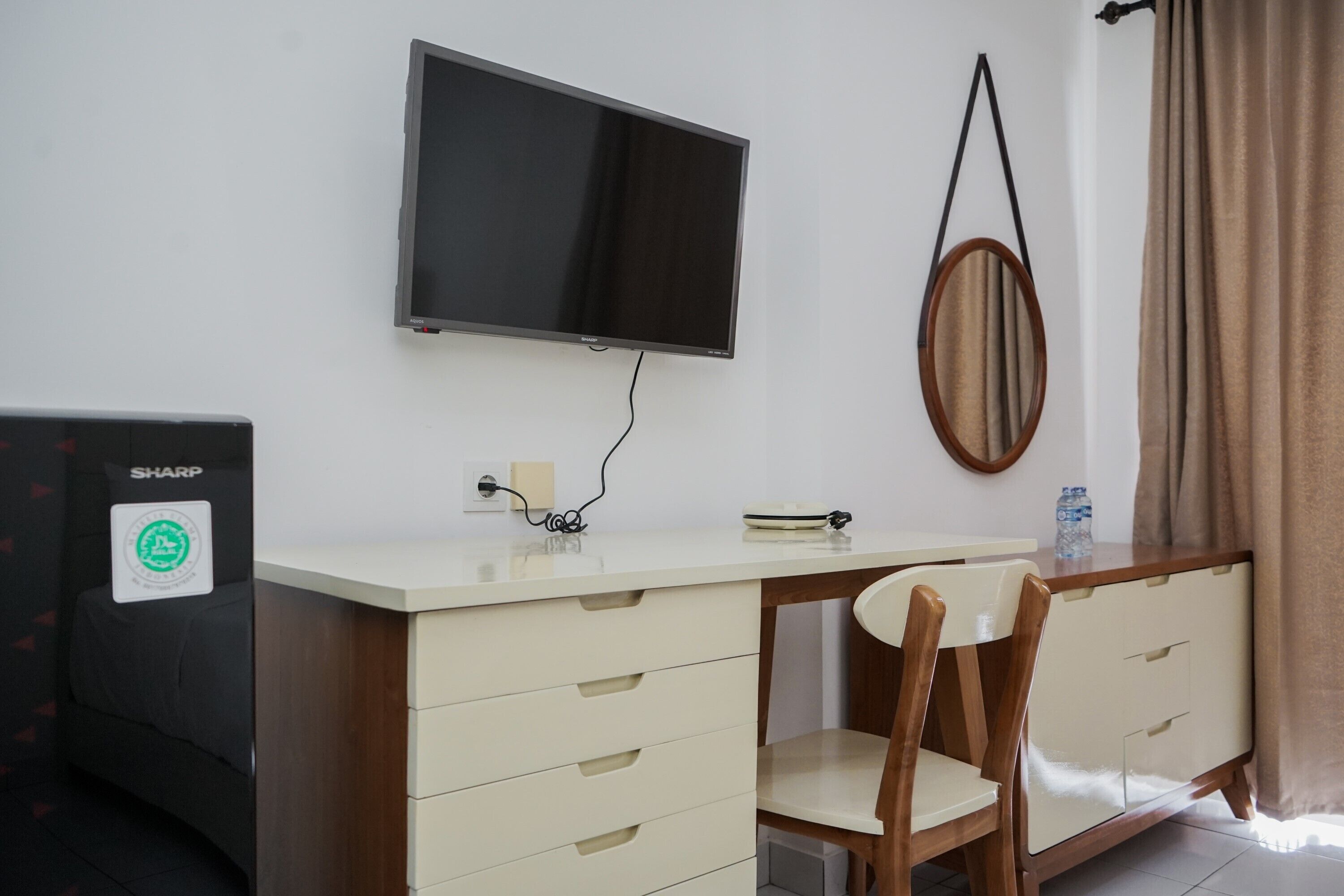 Habitación con televisión de pantalla plana montada en la pared, un escritorio con cajones, una silla, un monitor Sharp y un espejo.