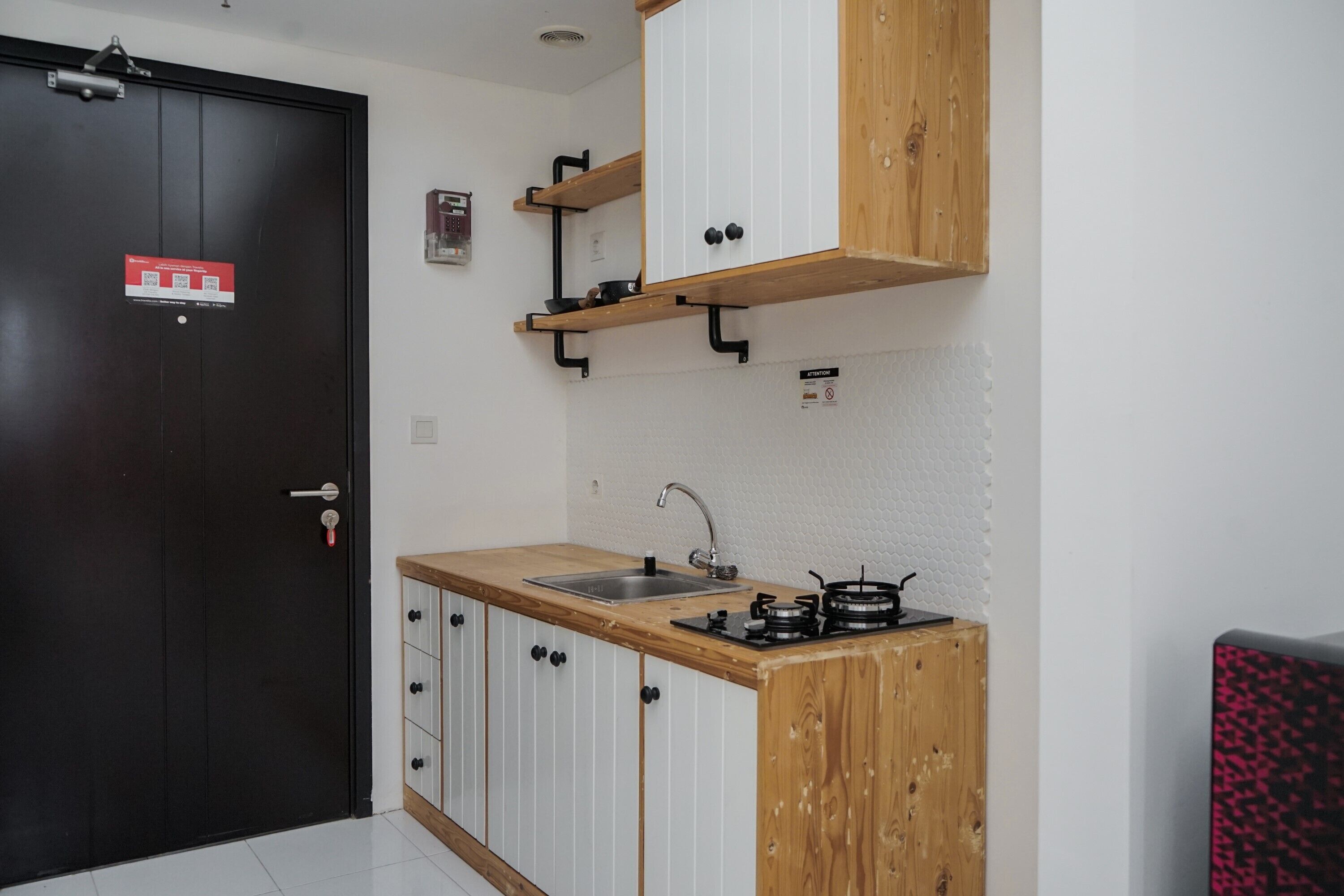 Une cuisine de taille réduite, dotée d’un plan de travail en bois, d’un évier et d’une cuisinière. On y trouve une porte noire avec une enseigne rouge, ainsi qu’un mur blanc à la texture particulière.