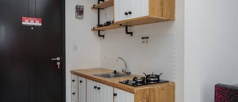 Une cuisine de taille réduite, dotée d’un plan de travail en bois, d’un évier et d’une cuisinière. On y trouve une porte noire avec une enseigne rouge, ainsi qu’un mur blanc à la texture particulière.