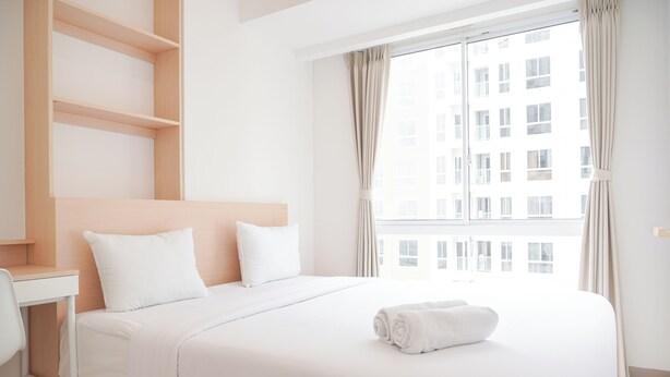 A hotel room with a bed, a desk, and a view of a building through the window. - Comfort Designed 2Br At Tokyo Riverside Pik 2 Apartment (Teluknaga)