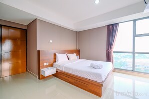 A modern hotel room with a large bed, wooden headboard, a nightstand, and a view of the city through large windows. - Comfort And Modern Look 3Br Apartment Royal Olive Residence (Jakarta)