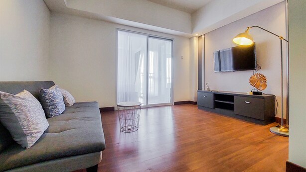 A modern living room with a grey sofa, a small round table, a flat-screen TV mounted on the wall, and a floor lamp. - Comfy And Minimalist 1Br At Tamansari Bintaro Mansion Apartment (South Tangerang)