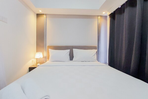A hotel room with a bed, a nightstand with a lamp, and a dark curtain. - Comfy And Minimalist 1Br At Tamansari Bintaro Mansion Apartment (South Tangerang)