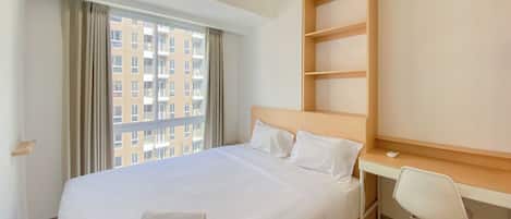 A hotel room with a large bed, a desk with a chair, and a view of a building through the window.