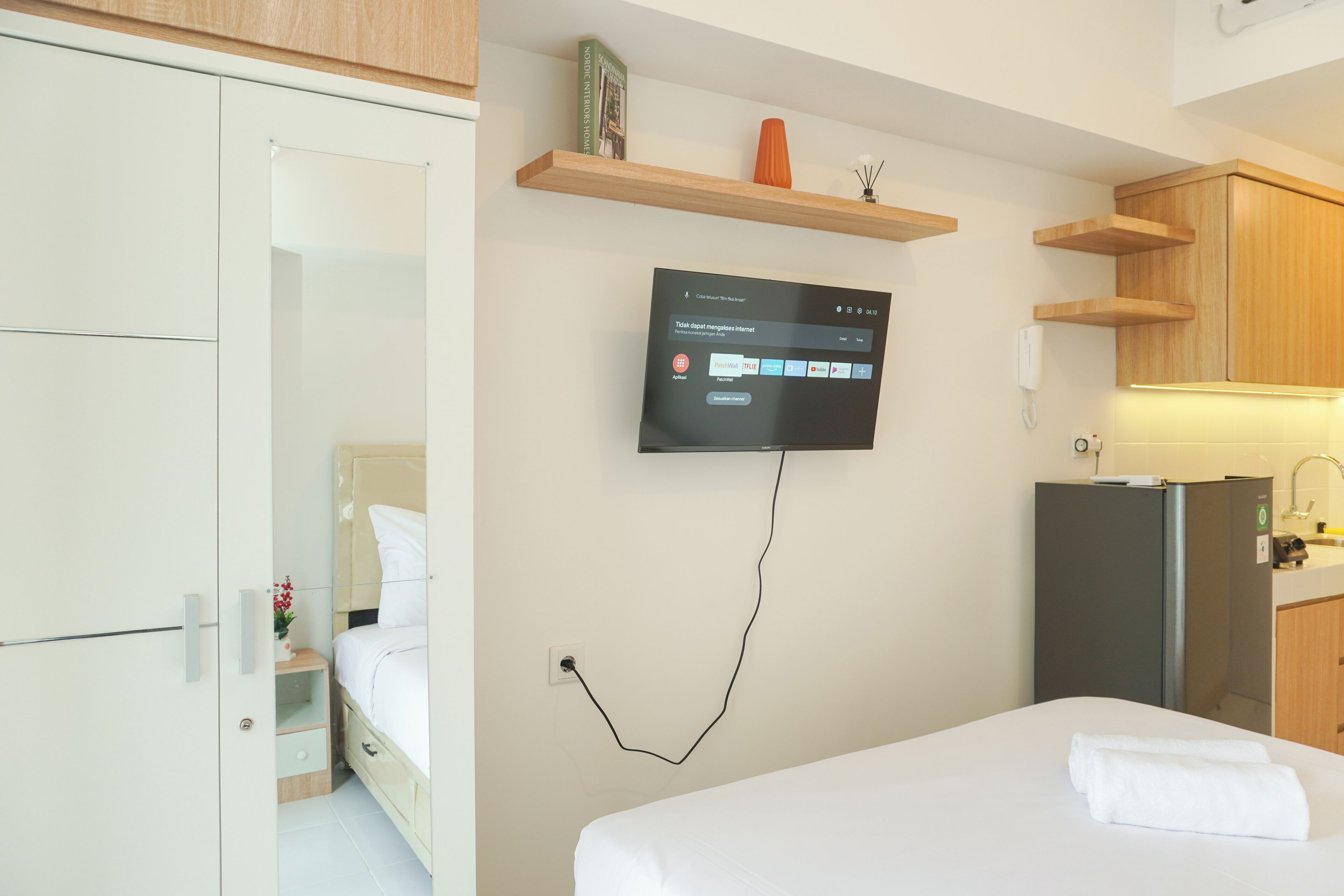 A hotel room with a bed, a TV mounted on the wall, a small table with a vase, and a refrigerator.