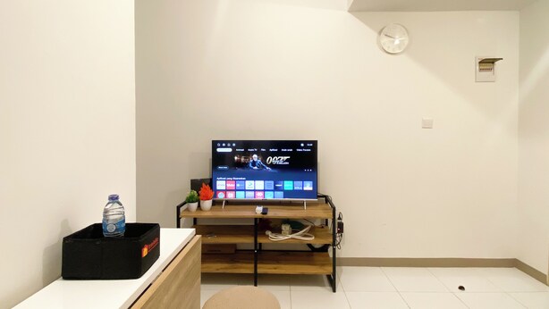 A room with a television on a wooden stand, a small table with a water bottle, a clock on the wall, and a power outlet. - Tidy And Homey 2Br At Tokyo Riverside Pik 2 Apartment (Teluknaga)