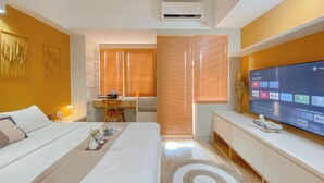 A modern hotel room with a bed, a desk, a TV, and a large window with blinds. - Good Studio Apartment LRT City Jatibening (Bekasi)