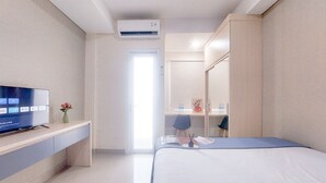 A modern hotel room with a flat-screen TV, a desk with a vase, a bed with a blue mat, a mirror, and a door leading to another room. - Good And Comfort Stay Studio Apartment At B Residence (Pagedangan)