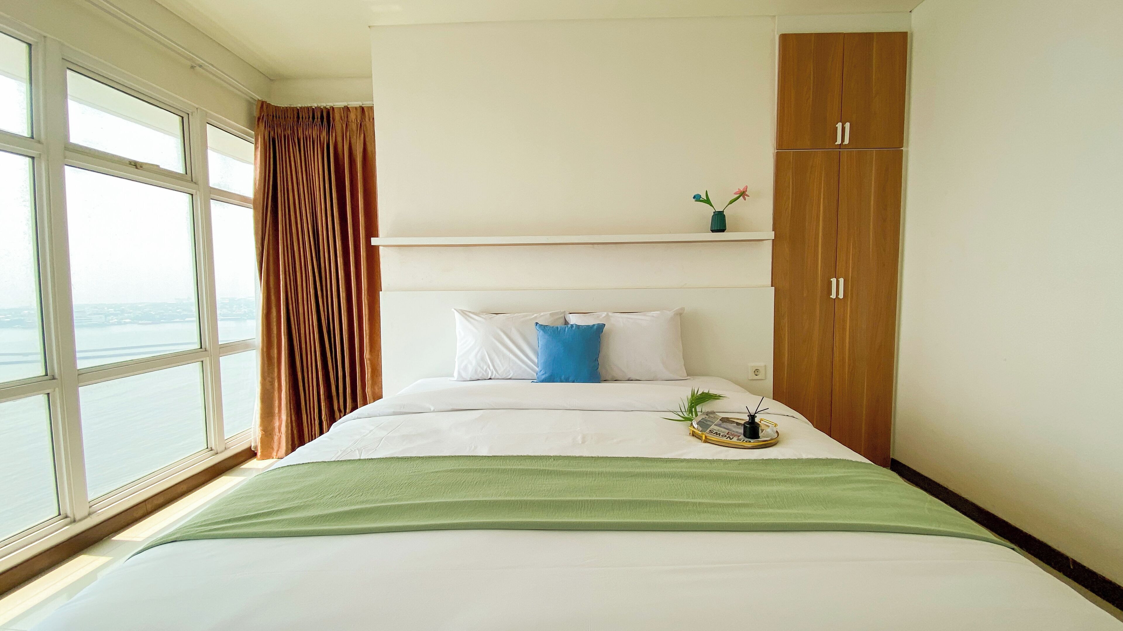 Une chambre d’hôtel avec un lit, une fenêtre avec des rideaux, une armoire en bois et une petite étagère sur laquelle se trouve un vase.