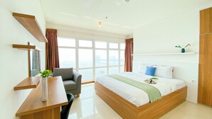 A modern hotel room with a large bed, a desk, and a view of the cityscape through large windows. - Best Homey And Spacious 3Br At Green Bay Condominium Apartment (Jakarta)