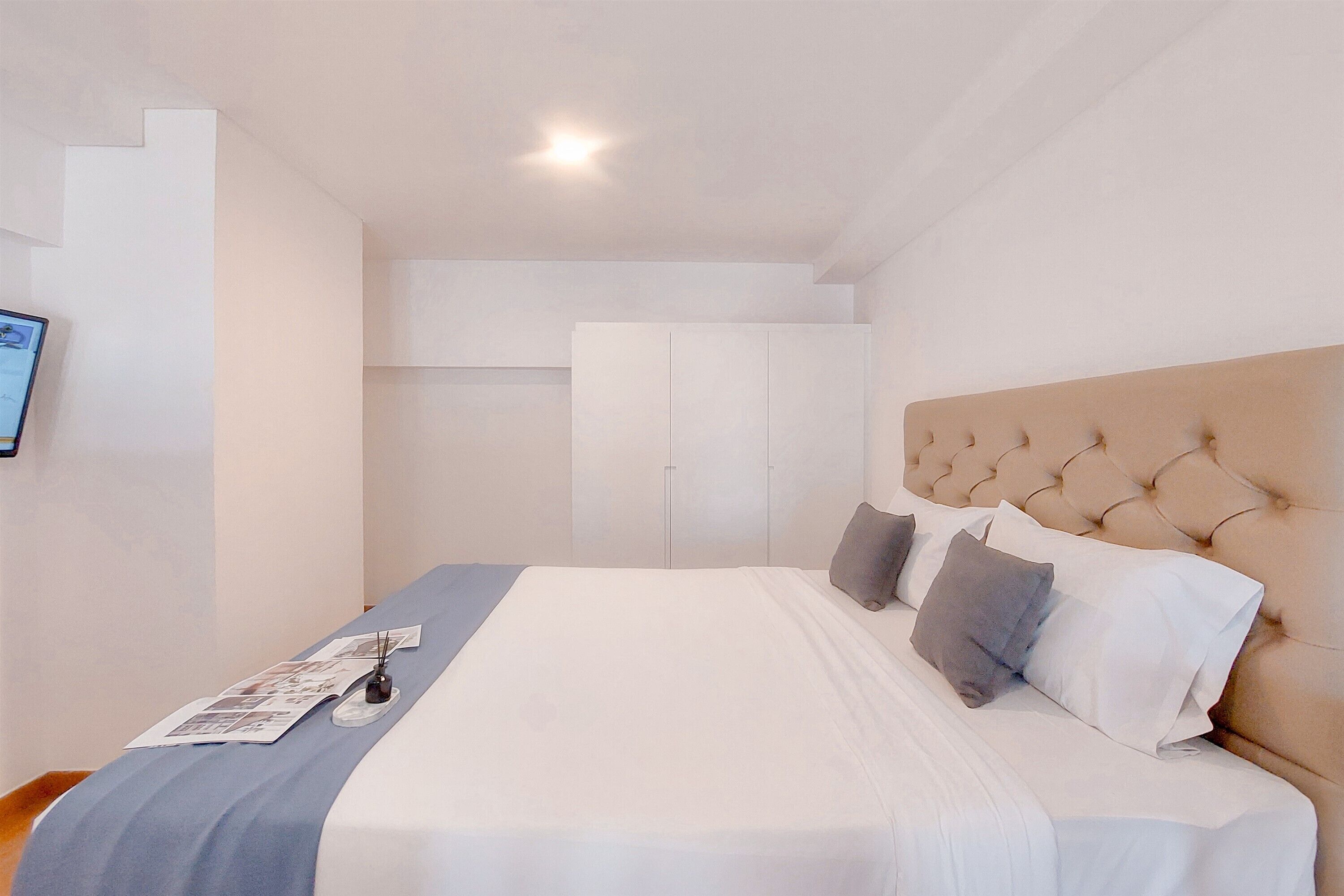 A bedroom with a large bed, a headboard, a bedside table with a lamp, and a wall-mounted shelf.