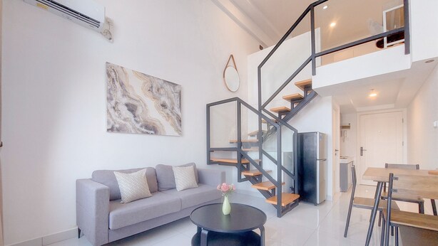 Una sala moderna con un sofá gris, una mesa redonda negra con un florero, una escalera con barandillas de vidrio y un comedor con mobiliario de madera.