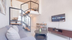 Una sala de estar moderna con un sofá gris, una mesa redonda, un televisor montado en la pared y una escalera con barandilla de vidrio.