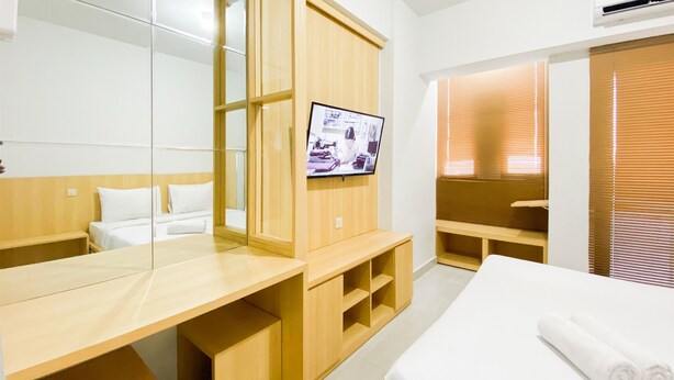 A hotel room with a large mirror, a TV mounted on the wall, a wooden desk, a bed with white linens, and a window with blinds. - Best Deal And Homey 1Br Without Living Room At The Alton Apartment (Semarang)