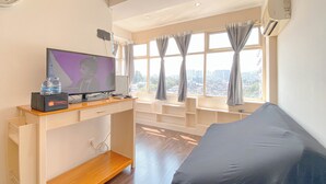 A modern hotel room with a large window, a flat-screen TV, a desk with a computer, and a covered sofa. - Spacious 1Br At Jarrdin Cihampelas Apartment (Bandung)
