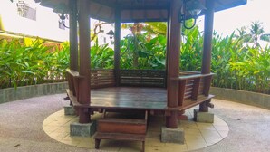 A wooden gazebo with a bench, surrounded by a circular tiled floor, and lush green plants in the background.