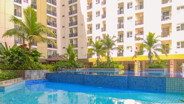 A swimming pool with blue tiles, surrounded by greenery and a multi-story building with balconies.