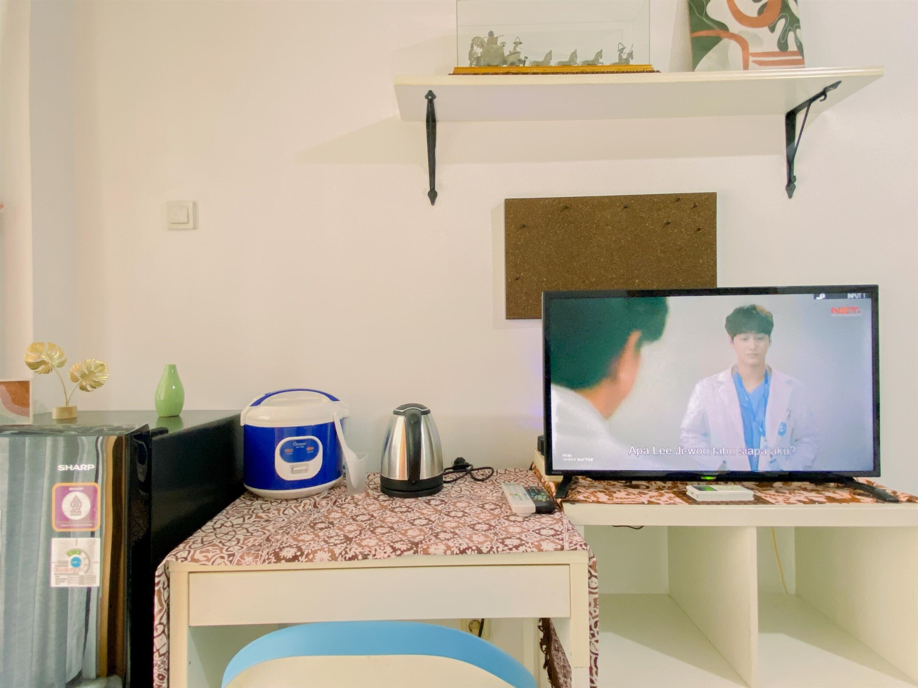 A room with a desk, a computer monitor displaying a show, a kettle, a blue container, and a small shelf with decorative items.