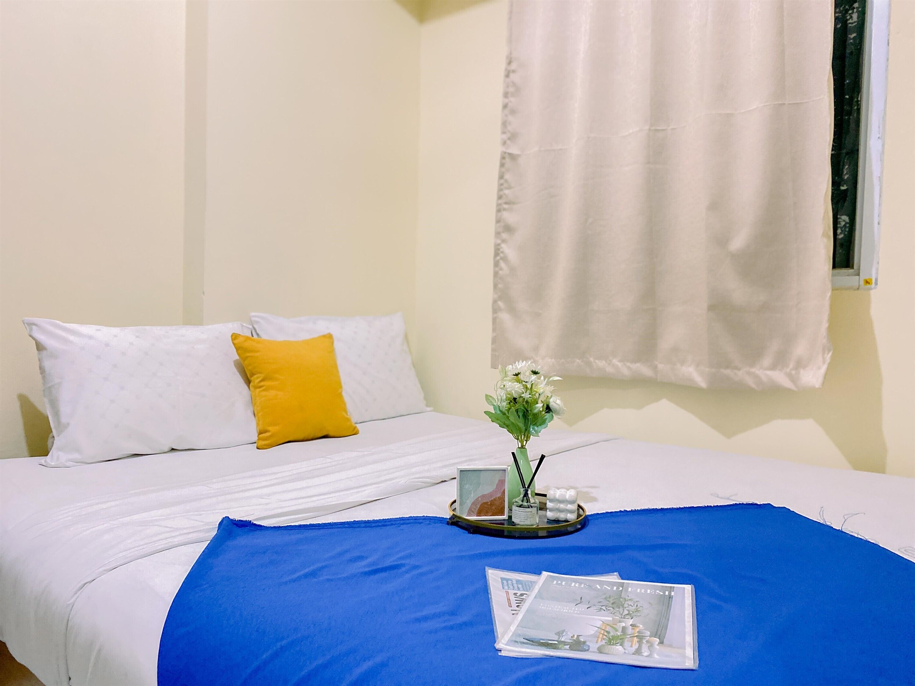 Una cama bien hecha con almohadas blancas y amarillas, una colcha azul y una mesita de noche con un florero y una revista.