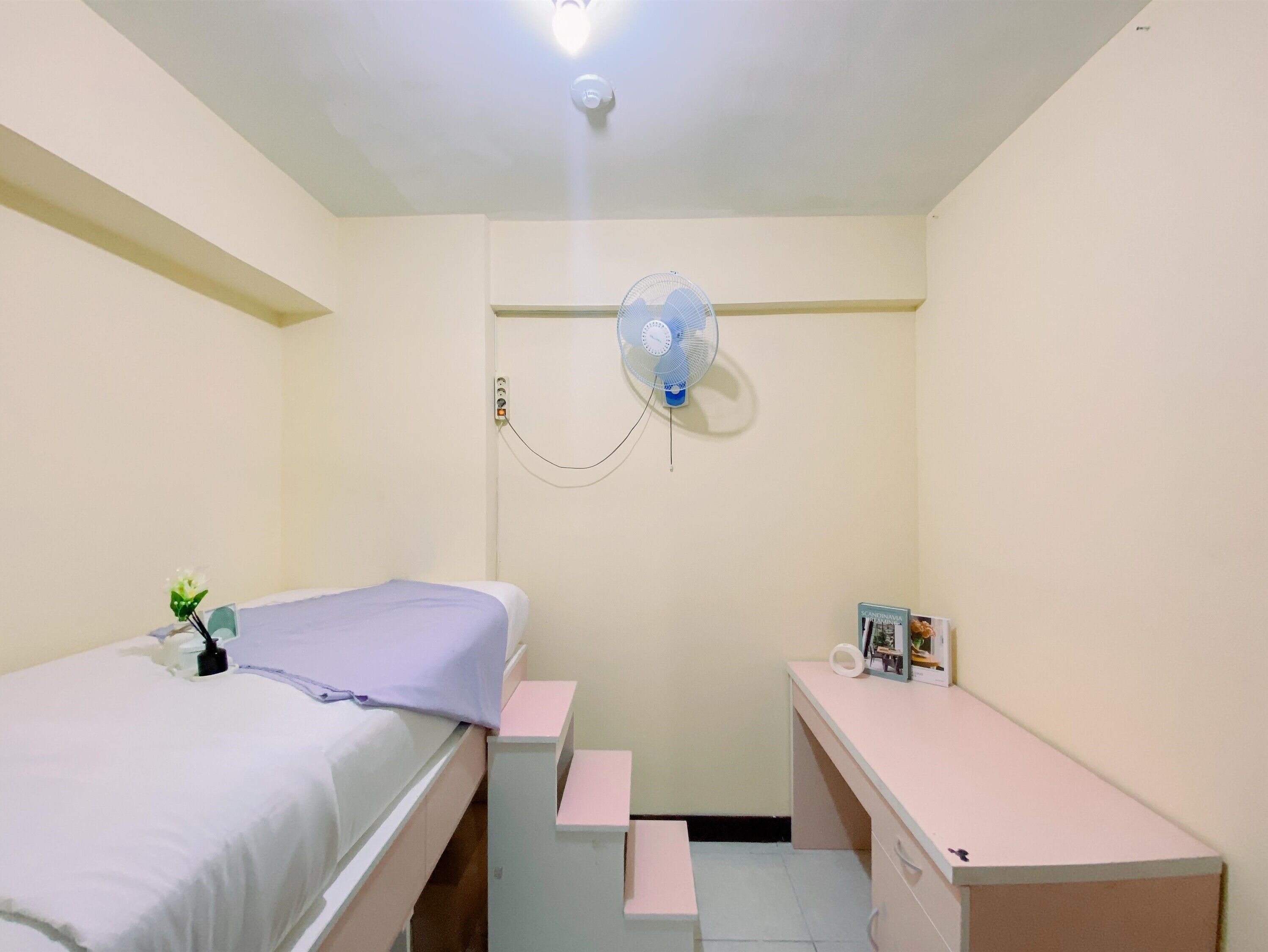 Et lille, godt oplyst værelse med seng, skrivebord og en stående ventilator.