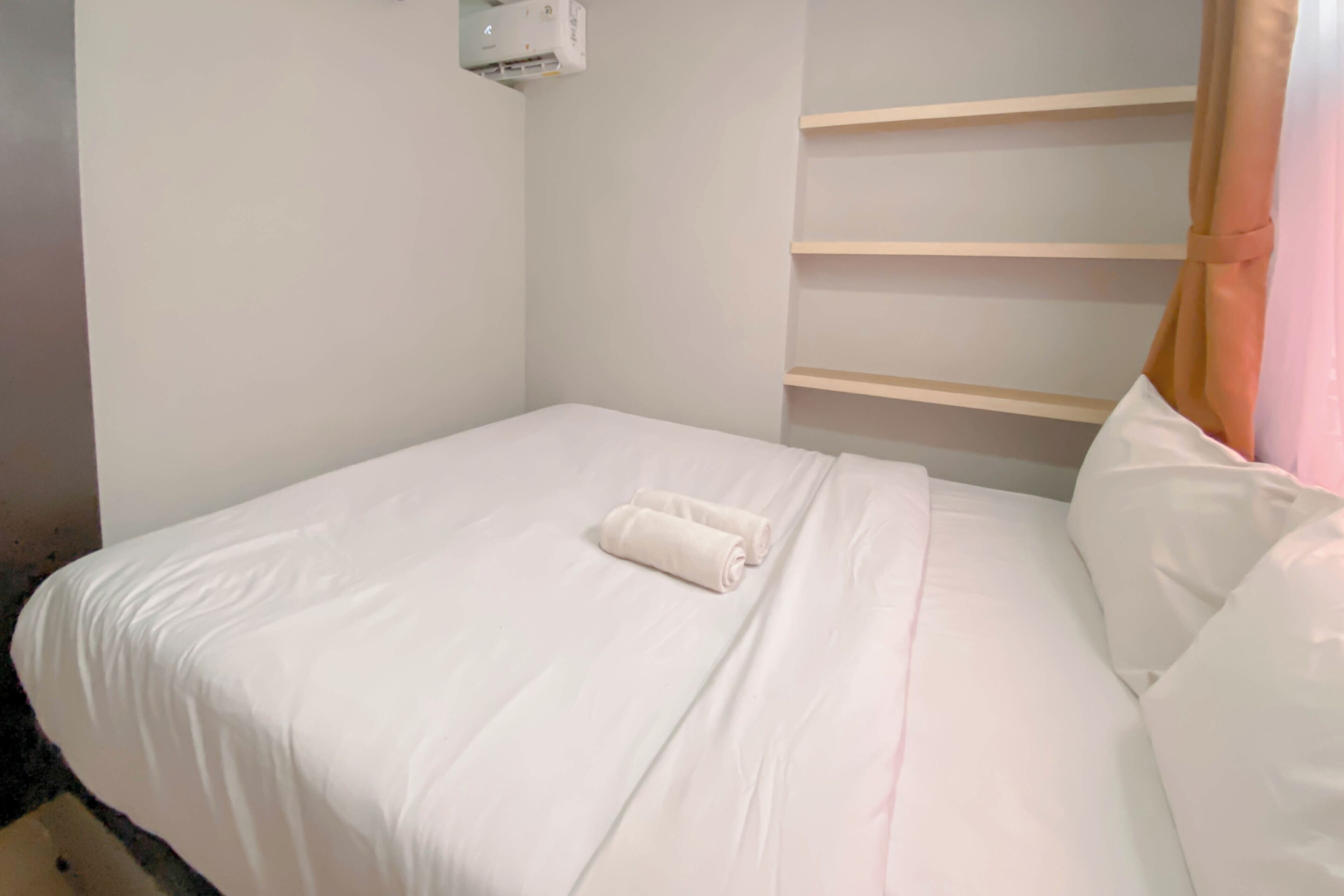A neatly made bed with white linens and a rolled towel, a wall-mounted air conditioner, and a wooden shelf unit against the wall.