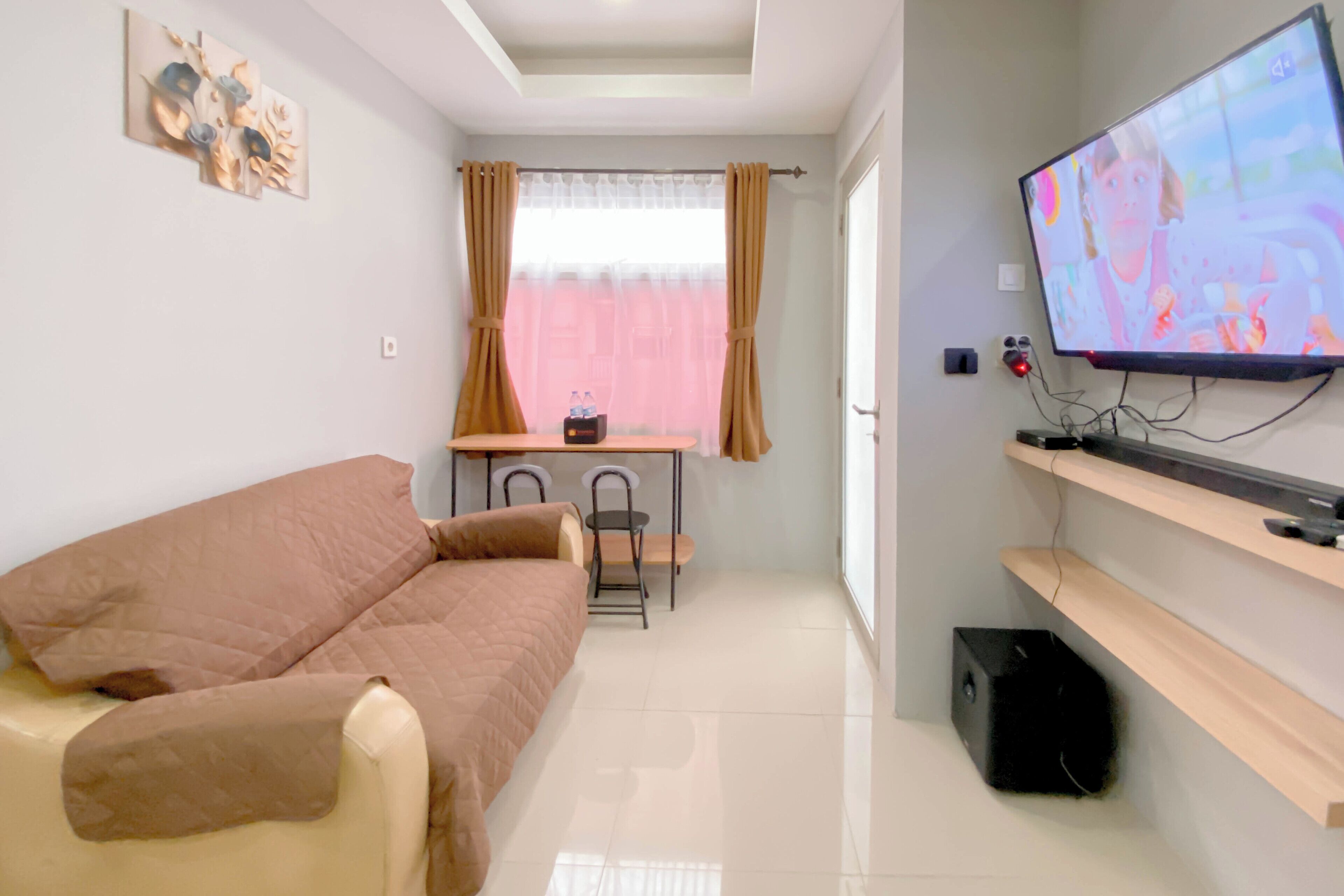 A compact room with a bed, a small table with chairs, a television mounted on the wall, and a window with curtains.