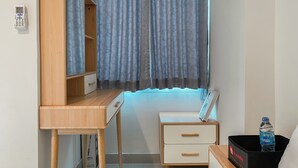 A bedroom with a wooden desk, a bedside table, a small drawer unit, and a window with curtains. - Minimalist And Cozy Studio Tamansari Iswara Apartment (Bekasi)