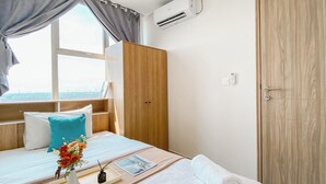 A hotel room with a bed, a wooden wardrobe, an air conditioning unit, and a view of the city through a window.