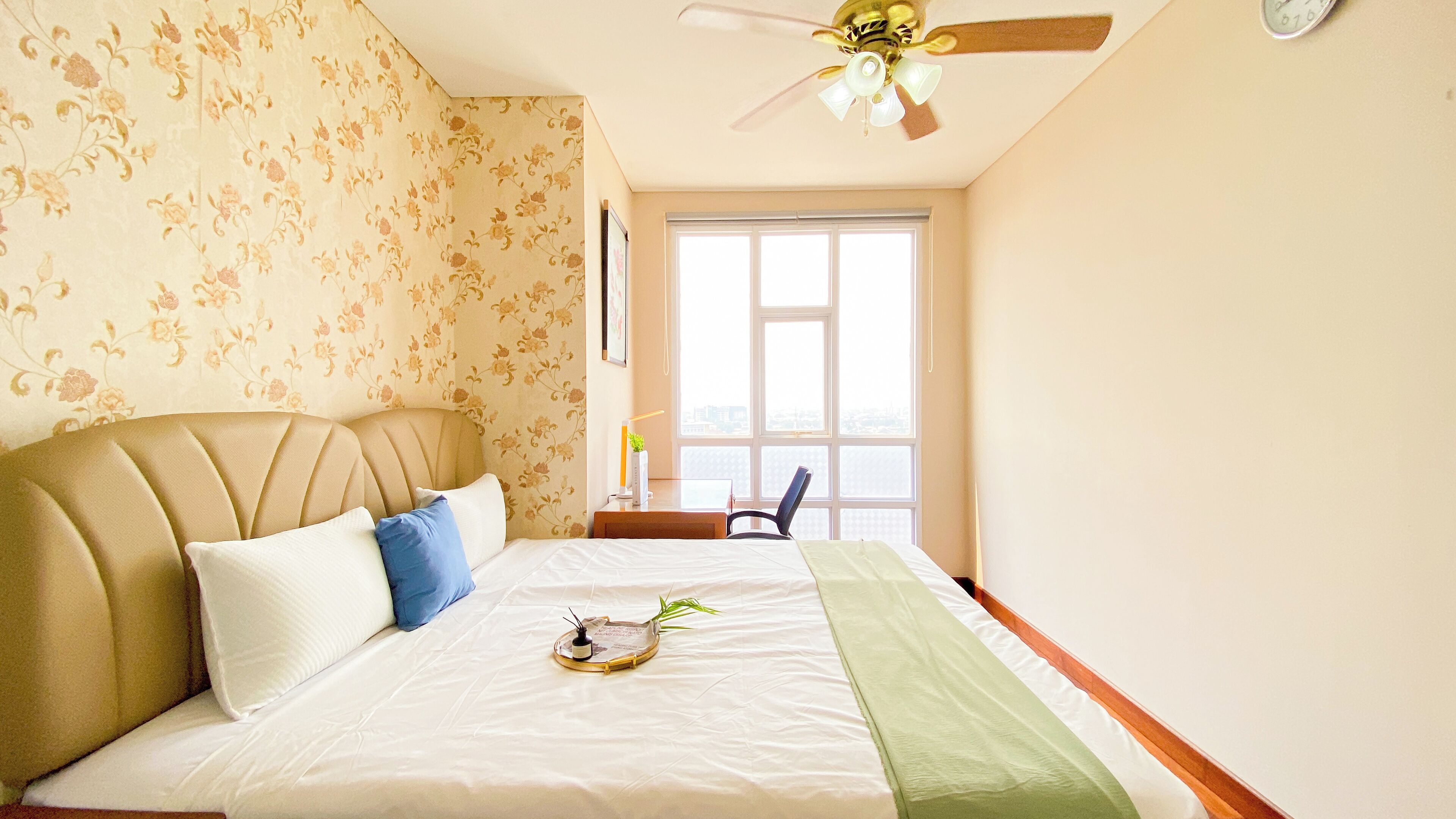 Un dormitorio con cama, ventilador de techo, ventana y papel tapiz floral.
