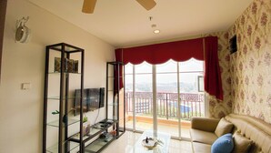 A living room with a glass TV stand, a sofa, and a balcony with a red curtain. - Modern Look 2Br At Lucky Tower Residence Apartment (Jakarta)