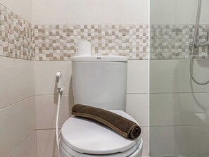 A white toilet with a brown towel on top, a shower head, and a tiled wall with a checkered pattern. - Spacious And Comfort 2Br Tamansari Iswara Apartment (Bekasi)