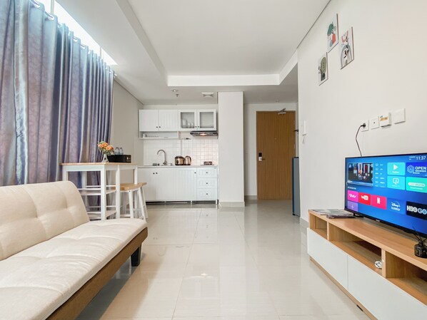 A modern living room with a sofa, a TV, and a kitchen in the background. - Spacious And Comfort 2Br Tamansari Iswara Apartment (Bekasi)