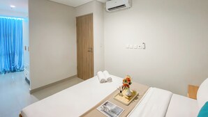 A hotel room with a bed, a bedside table with a tray, a wall-mounted air conditioner, a door, and a curtain.