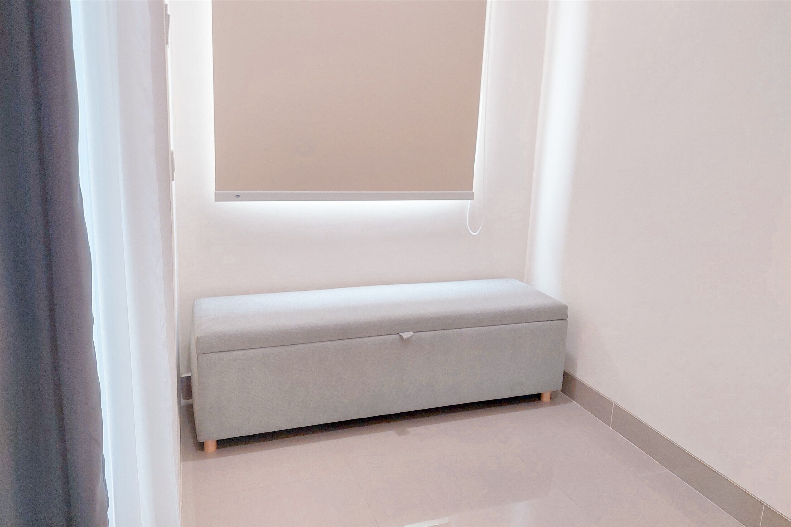 A small, light-colored bench in a corner of a room with a window.