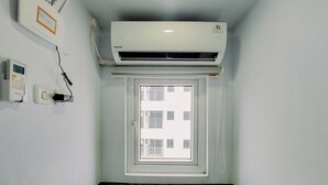 Una habitación con aire acondicionado integrado, una ventana con persianas, un termostato fijado a la pared y piso de madera.