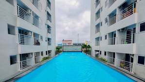 Una piscina flanqueada por edificios de dos plantas con balcones, un cielo despejado y una torre en la distancia.