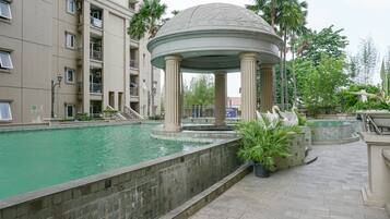 Zona de piscina con un pabellón, palmeras y un edificio de varios pisos al fondo.