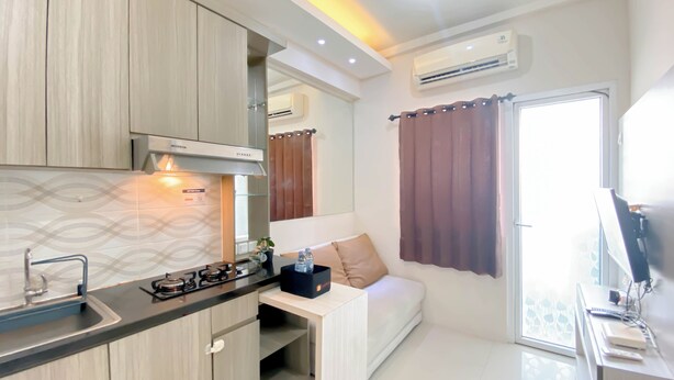 A modern kitchen with a gas stove, sink, and wooden cabinets. There is a small dining area with a white table and a sofa. An air conditioner is mounted on the wall, and a window with curtains is visible.