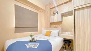 A hotel room with a bed, a small stool, a vanity with a mirror, and a wall-mounted shelf with decorative items.