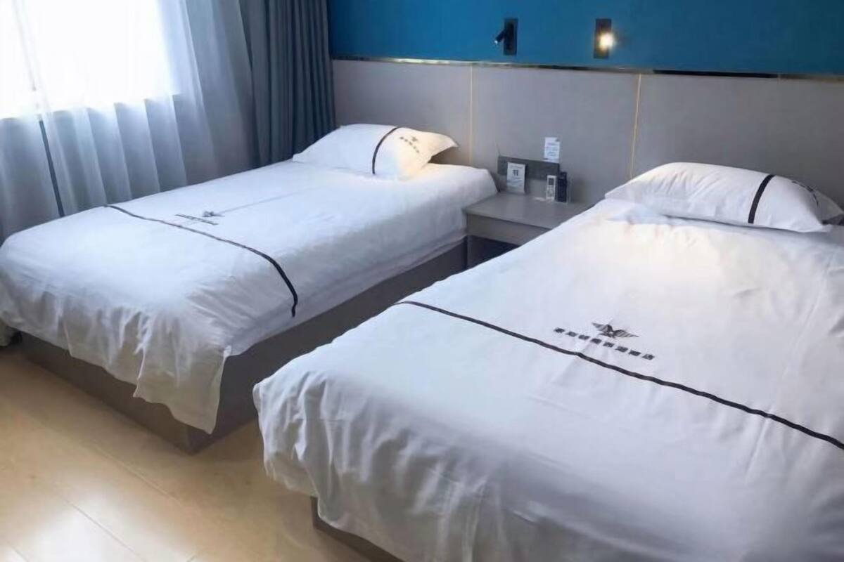 Two single beds with white bedding and a small bedside table with a lamp, in a room with a blue accent wall and a window with curtains.