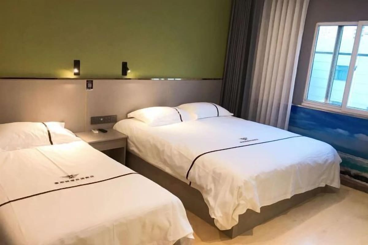 Quarto de hotel com duas camas, uma mesa de cabeceira, uma luminária na parede e uma janela com cortinas.