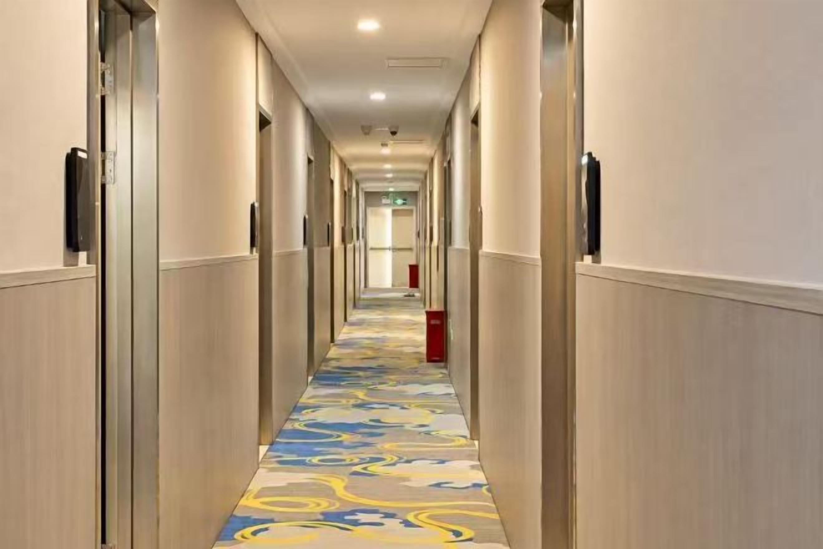 Un couloir d’hôtel avec des portes des deux côtés, une moquette à motifs et une poubelle rouge au centre.