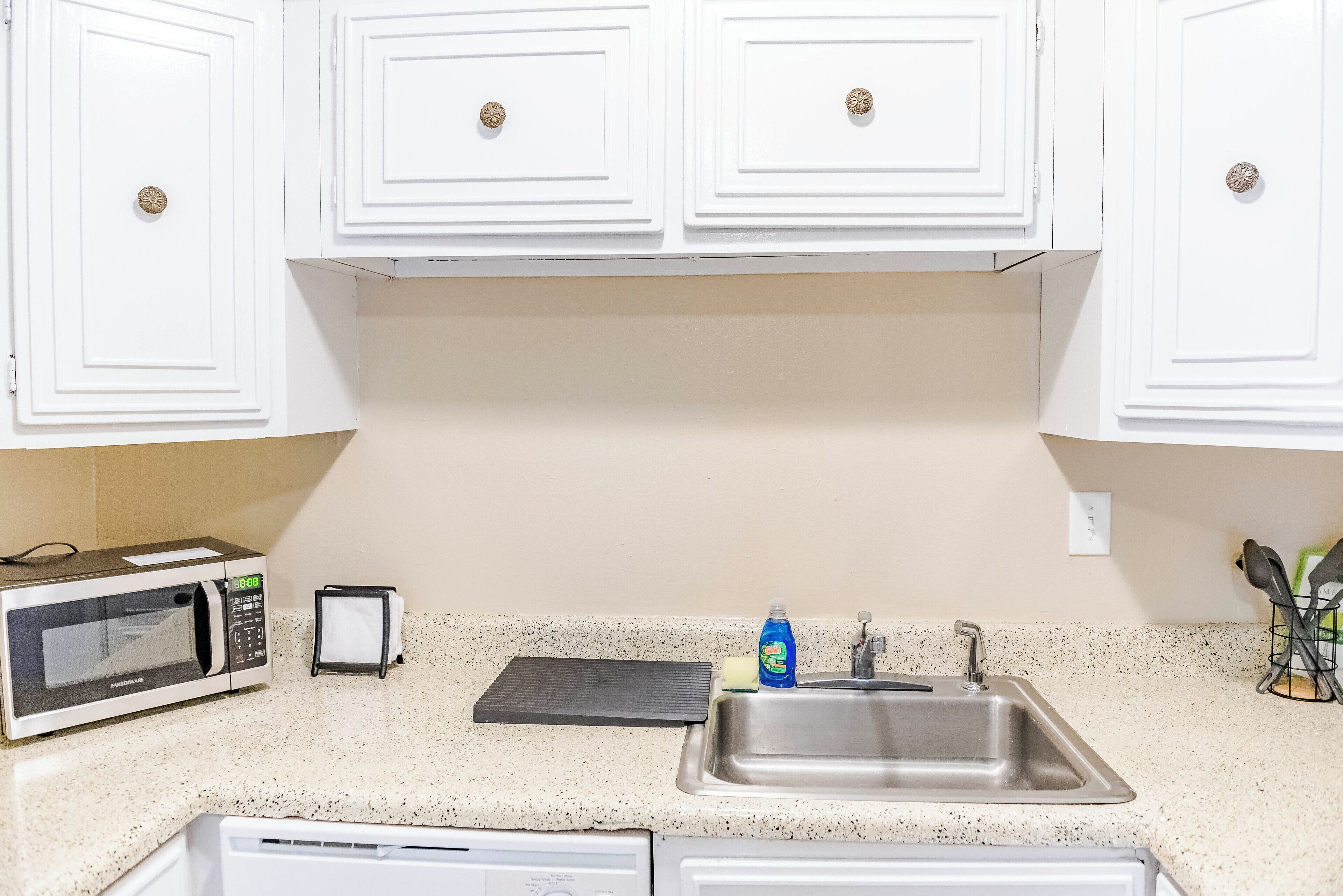 Una cocina con armarios blancos, un lavamanos de acero inoxidable, un microondas, un escurreplatos y una botella de detergente.