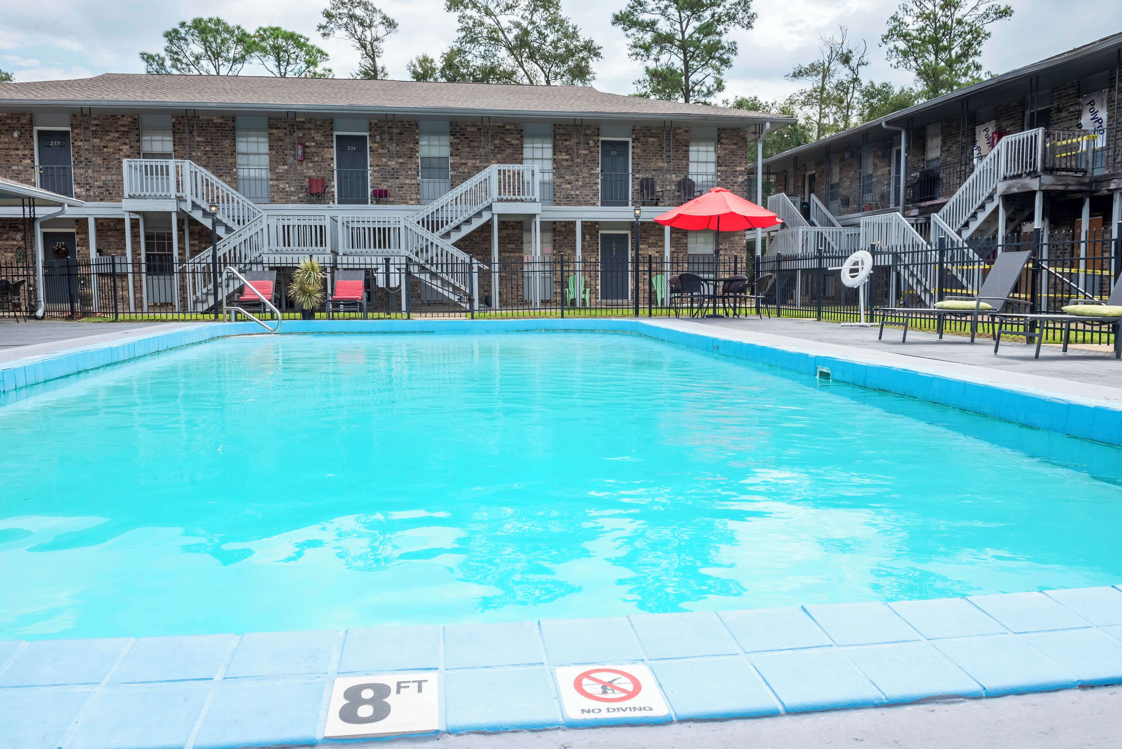 Una piscina con un cartel de prohibición de buceo, un indicador de profundidad de 2.5 metros y una zona cubierta con sombrillas rojas.